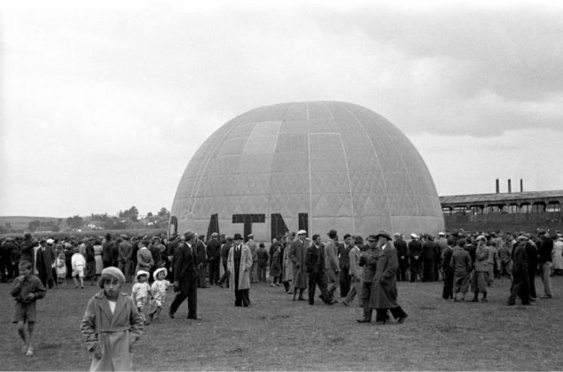 Pierwszy lot balonu „Sanok” Autor: Stanisław Potocki Czas wykonania: 21.06.1936 r. Fotografia przedstawia balon „Sanok” na sanockich błoniach nad Sanem utrwalony w trakcie napełniania. Wokół balonu zgromadzeni widzowie. Z prawej, w tle, widoczne zabudowania Fabryki Gumy.