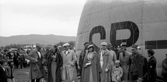 Pierwszy lot balonu „Sanok" Autor: Stanisław Potocki, 21.06.1936 r. Fotografia przedstawia balon „Sanok” na sanockich błoniach nad Sanem utrwalony w trakcie napełniania. Wokół balonu zgromadzeni widzowie. Przed balonem grupa osób. Trzeci z lewej (w jasnym kaszkiecie) Oskar Schmidt – założyciel i dyrektor Fabryki Gumy oraz prezes sanockiego Klubu Balonowego. Obok, prawdopodobnie, jego żona.